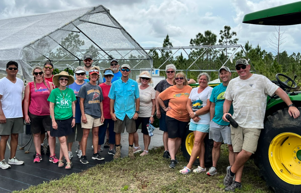 A group of 16 standing outside by a tractor and covered structures